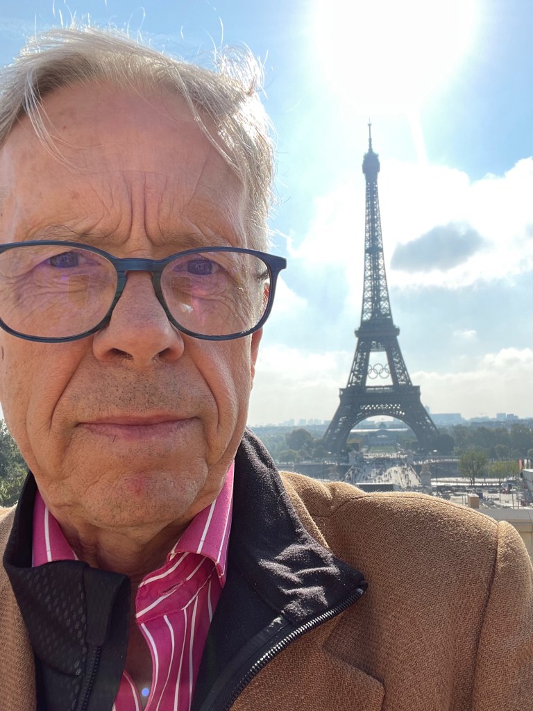 Un homme en gros plan avec des lunettes, se tenant devant la tour Eiffel sous un ciel dégagé.